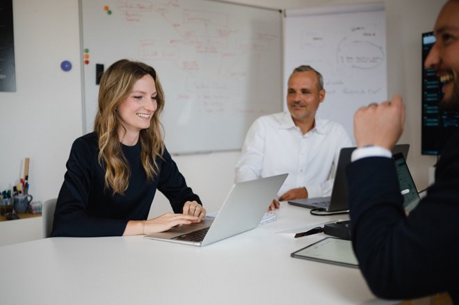 „Dynamische Team-Szene bei M2: Drei Personen im Austausch an einem runden Tisch mit Whiteboards und Laptops – modernes Arbeitsumfeld und kollegiale Atmosphäre.“